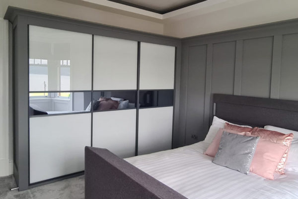 A modern bedroom features a gray fitted wardrobe with sliding doors and a bed with white linens and pink accent pillows.