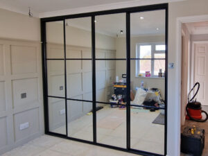 Modern room with beige panel walls features a large, black sliding wardrobe. Reflective surfaces show scattered renovation tools and natural light from a window.
