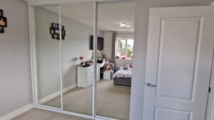 A bedroom with white sliding wardrobe doors reflects a well-organized space. A bed, nightstand, and wall art are visible, creating a calm, tidy ambience.