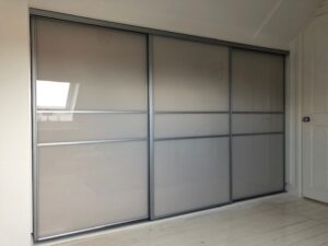 Sleek frosted grey glass sliding wardrobe doors with silver frames span across a light, minimalist room, reflecting soft natural light from a nearby window.