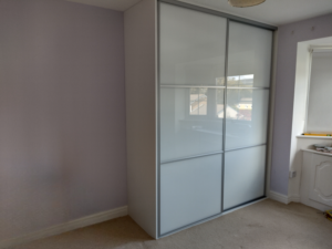 White sliding wardrobe in a minimalist room with light purple walls and beige carpet. A window seat with a few items is visible beside the wardrobe.