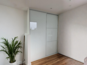 Minimalist room corner with white walls, a wooden floor, and a pure white sliding door wardrobe. A green potted plant adds a touch of nature.