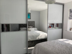 Modern bedroom with a plush gray headboard and white bedding, reflected in a white sliding wardrobe with mirrors. A bright, minimalist ambiance with soft sunlight.