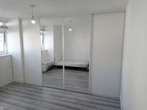 Bedroom with a white sliding wardrobe reflecting a bed and windows. The space is bright, with gray wood flooring and white walls, creating a clean, modern feel.