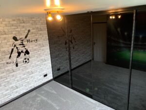 Room with a gray brick wall featuring a soccer player silhouette and "Harry" text. A 4 Door Black Sliding Wardrobe reflects a football stadium mural, under warm ceiling lights.