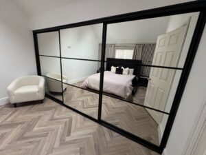 Modern bedroom with wood floor, a 3 door black sliding wardrobe, beige armchair, and neatly made bed, creating a cozy ambiance.