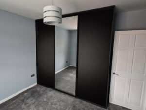 Stylish black wardrobe with sliding doors, including a large mirror, complementing a minimalist bedroom with grey carpeting.
