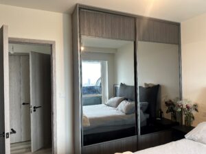 Modern bedroom with a large 2 door woodwork wardrobe reflecting a bed with a dark headboard, gray bedding, and a small teddy bear. With Fresh flowers on a nightstand.